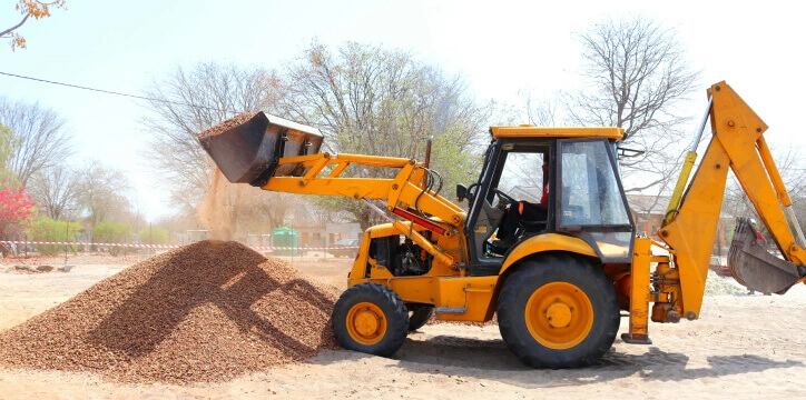Construction fleet management – a yellow backhoe loader moves gravel at a worksite with trees in the background.
