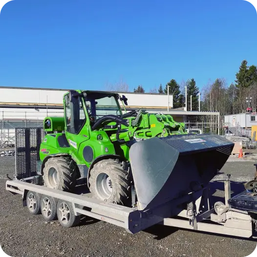 Cargadora compacta verde sobre un remolque en una obra, representando el equipo rastreado con Mapon por el cliente Tampereen Lainaväline.