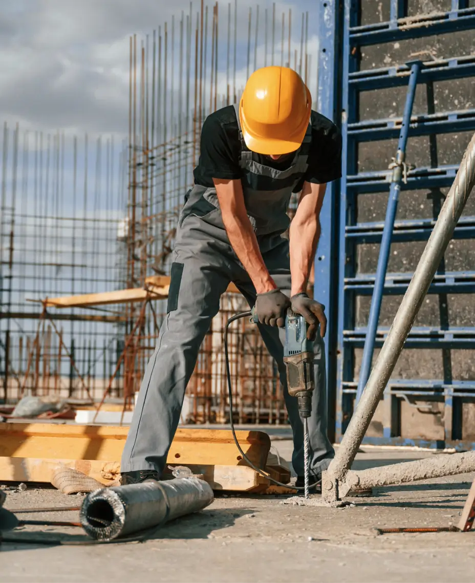 Trabajador de la construcción con casco amarillo y mono gris usando un taladro sobre una superficie de hormigón en una obra.