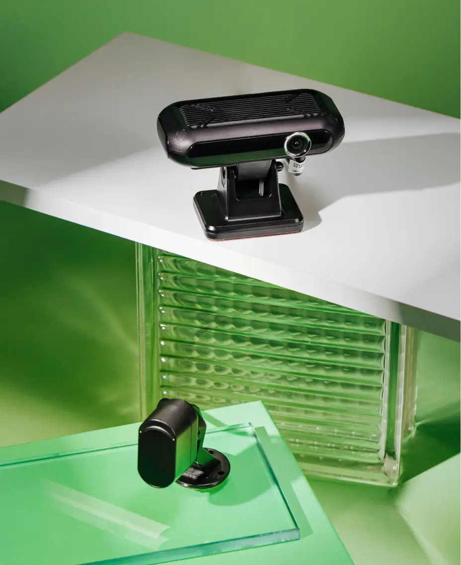 Two black Mapon fleet cameras with mounts, placed on glass and a white surface against a green background.