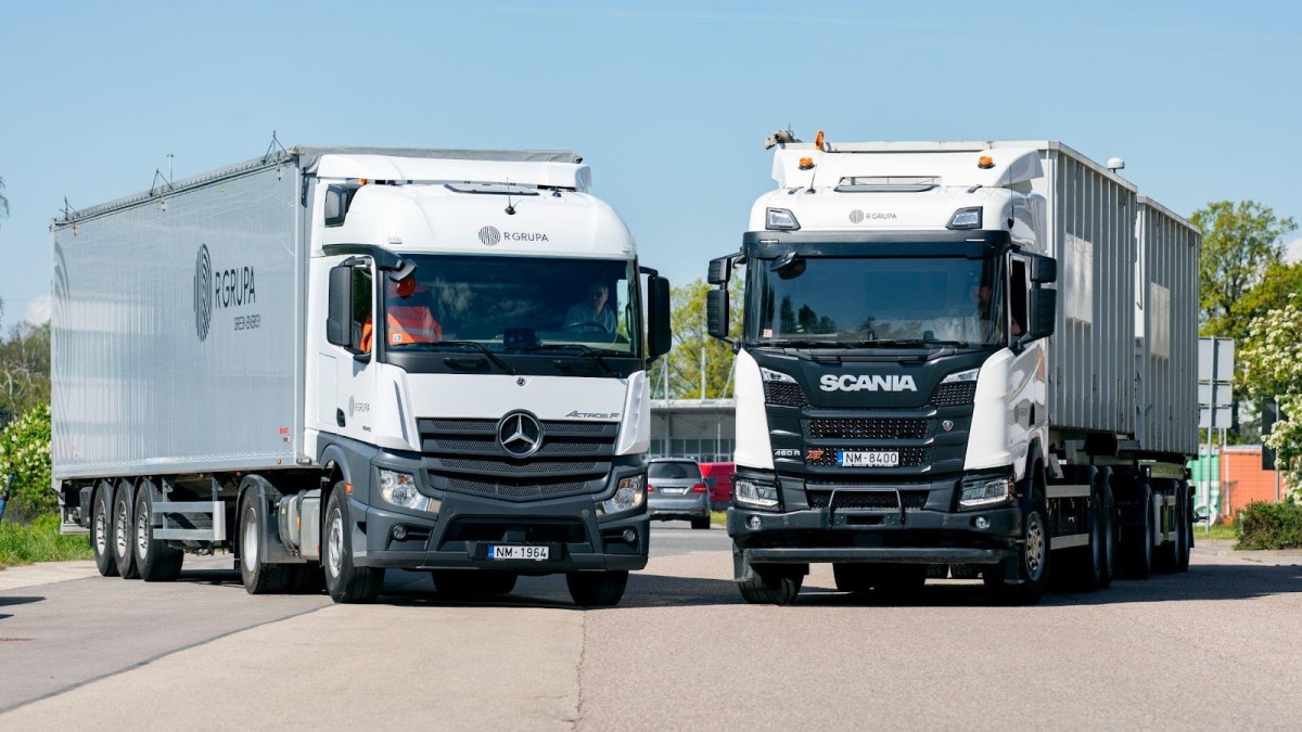 Divas baltas kravas automašīnas ar R GRUPA logotipiem stāv uz ceļa leņķī viena pret otru.