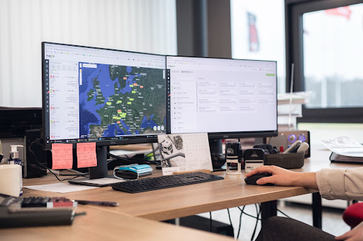 Person arbejder ved et skrivebord med Mapon-platformen åbnet på to skærme, der viser køretøjsplaceringer i Europa og flådedata.