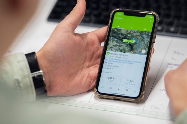 Person holder en smartphone med Mapons mobilapp åben, der viser en rute og køretøjets placering på kortet.